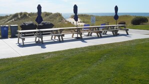 Een picknickplaats aan het strand met banken en parasols, een bordje dat toegang tot het strand aangeeft, en uitzicht op de oceaan.