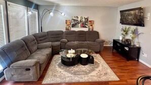 A living room with a sectional sofa, a glass coffee table, and a patterned rug. There are wall-mounted lights and a flat-screen TV.