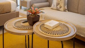 A modern hotel room with a sofa, two small round tables, a potted plant, and a large window with blinds. - Sleek and Stylish 1br in Mall Of Asia Complex (Pasay)
