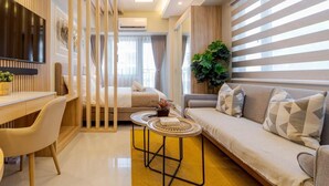 A modern hotel room with a sofa, a dining area with a table, a TV mounted on the wall, and a large window with blinds. - Sleek and Stylish 1br in Mall Of Asia Complex (Pasay)