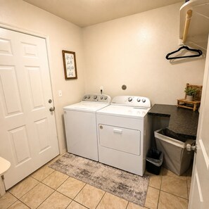 A laundry room with a washer and dryer, a trash can, and a small shelf with a plant.