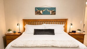 A neatly made bed with a white duvet and a dark pillow, flanked by two bedside tables with lamps and clocks, in a room with a simple wooden headboard and a wall-mounted artwork.