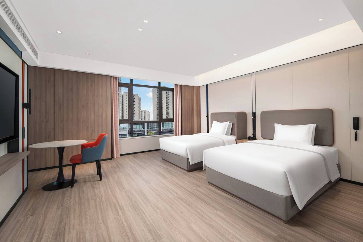 Habitación de hotel moderna con dos camas, una mesita con una silla y un ventanal con vistas a la ciudad.