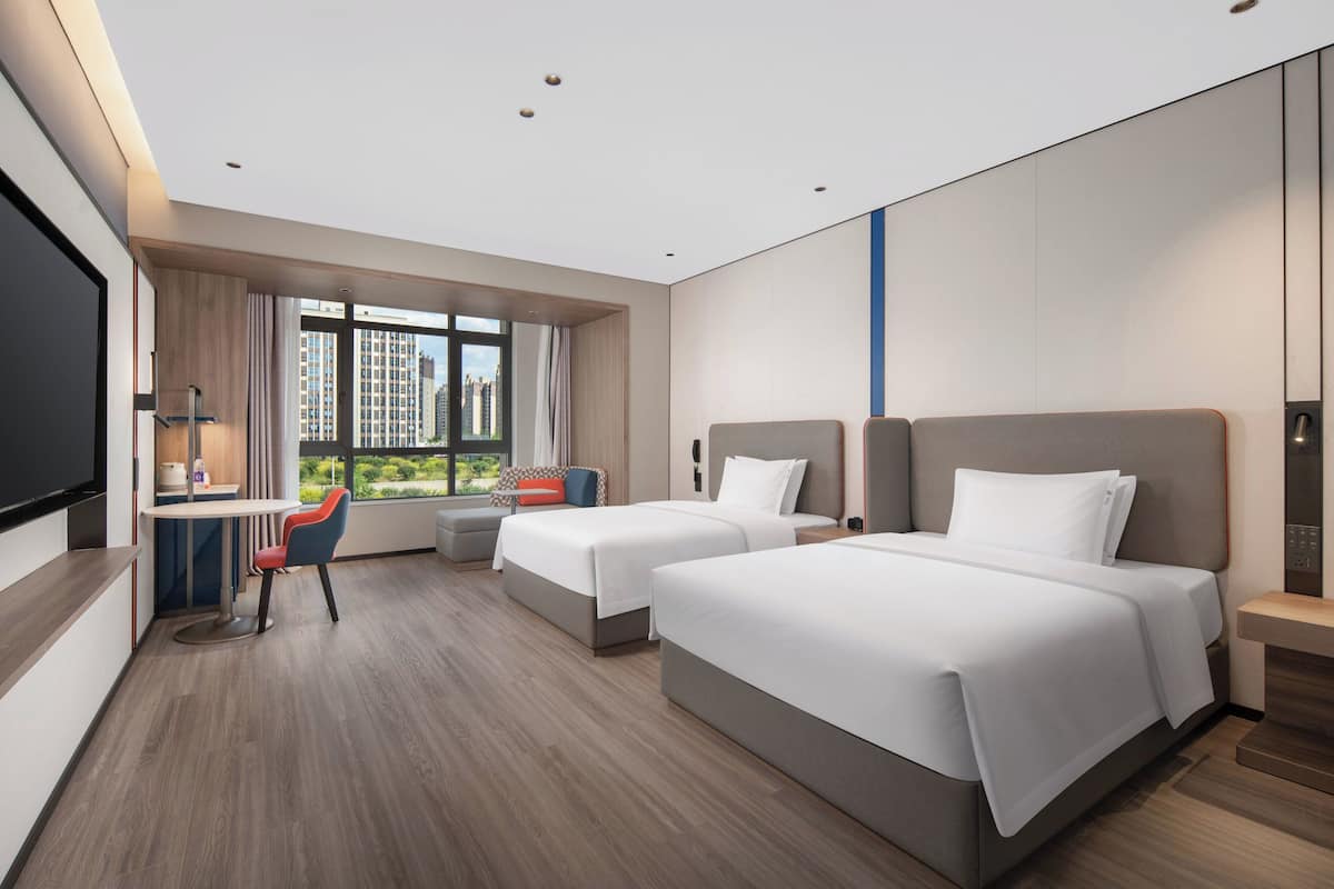Una habitación de hotel moderna con dos camas, un escritorio con silla, televisión y un ventanal con vistas a la ciudad.
