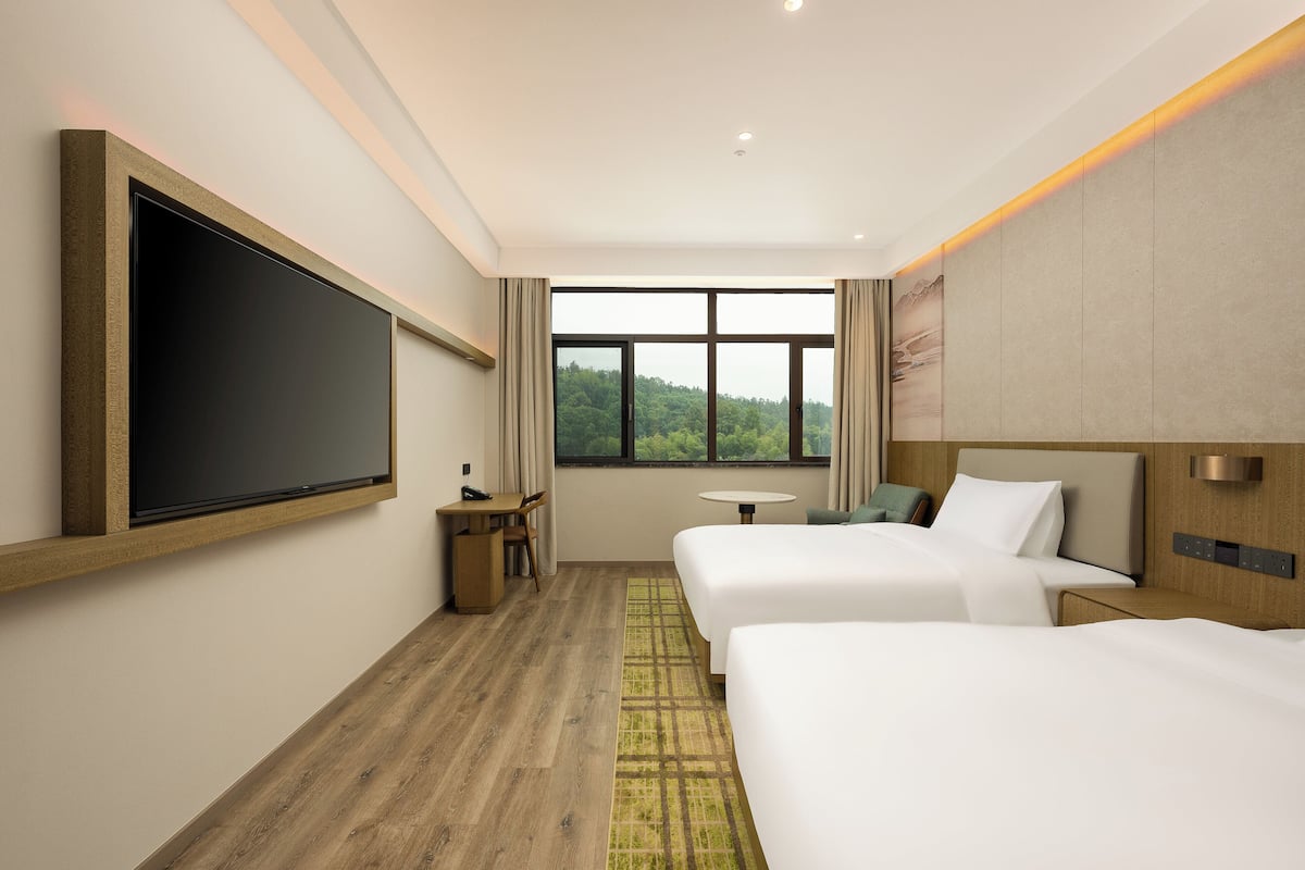 A hotel room with two beds, a desk, a chair, a small table, and a large window with a view of greenery.