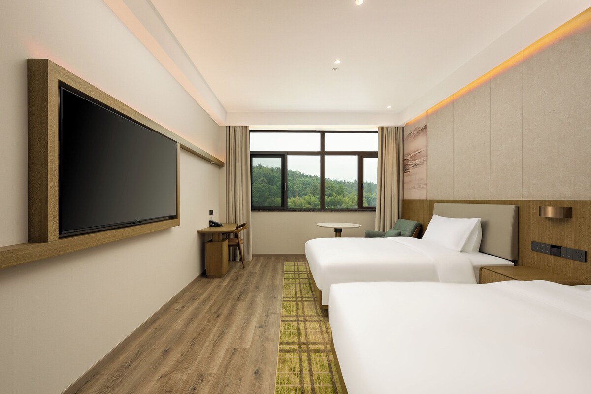 Habitación de hotel con dos camas, un escritorio, una silla, una mesita y un ventanal grande con vistas al verdor.