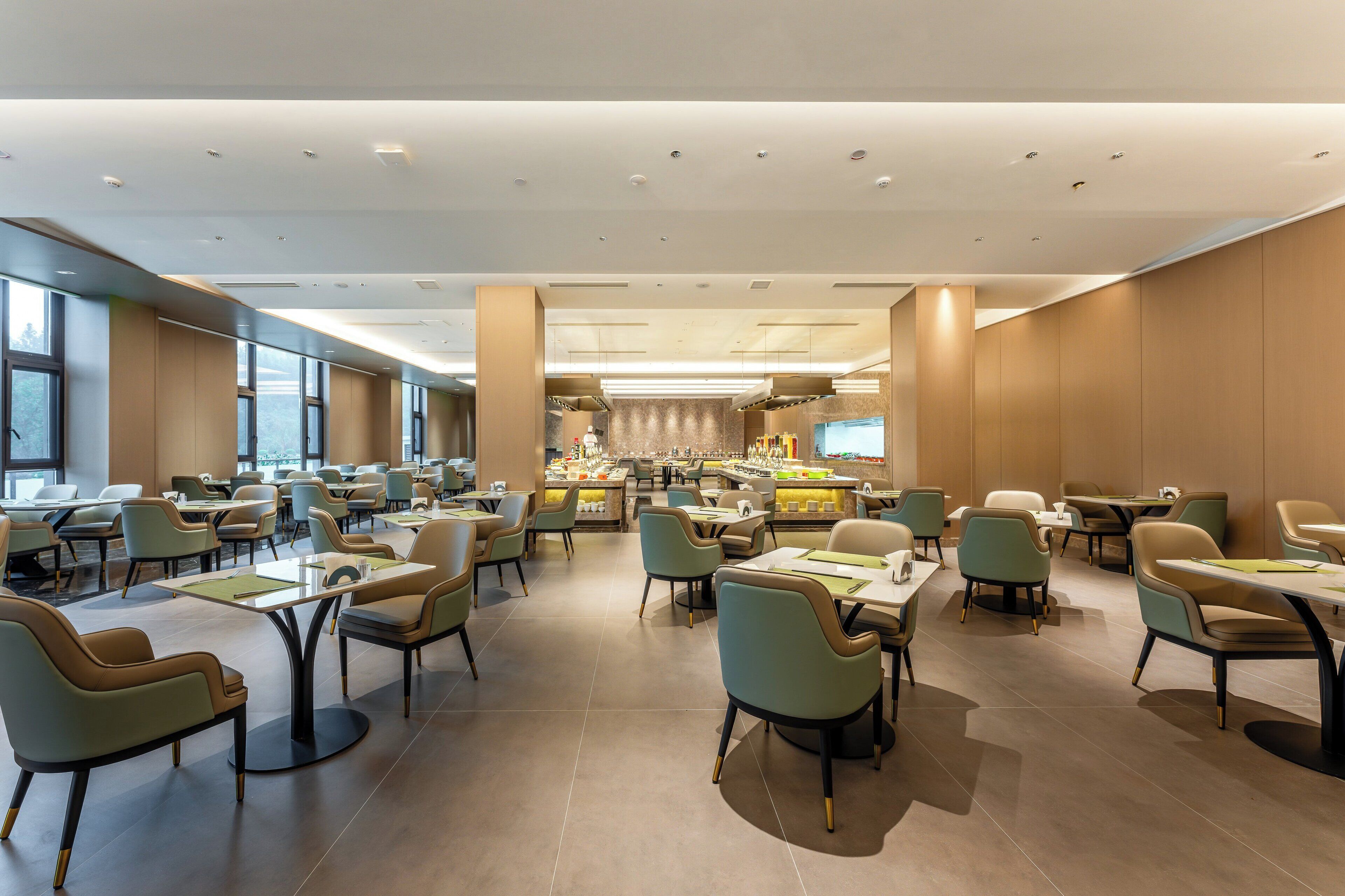 A spacious hotel dining area with multiple tables and chairs, a central bar, and large windows providing natural light.