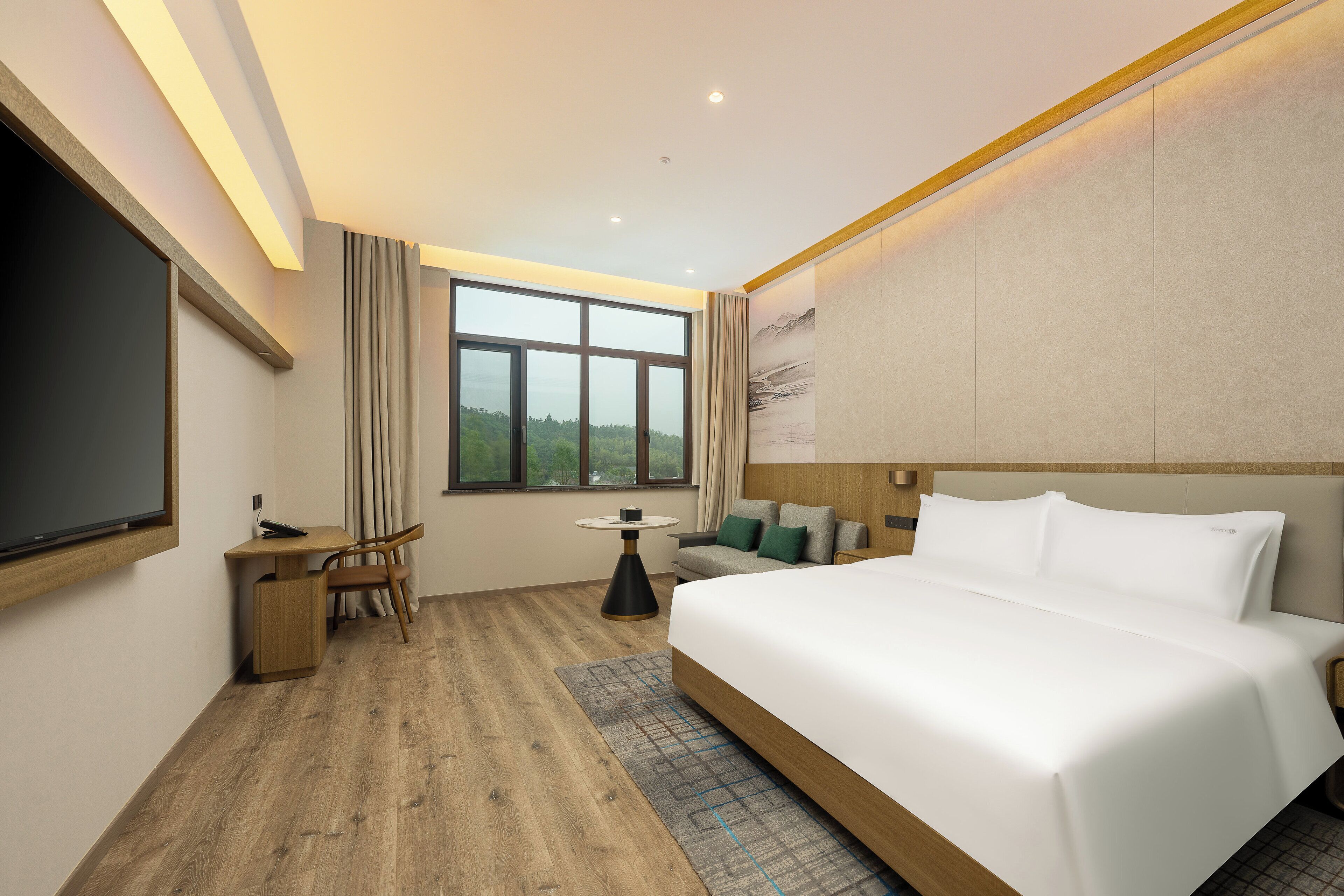 Una habitación de hotel moderna con una cama grande, un escritorio con silla, una mesita pequeña y un amplio ventanal con vistas a zonas verdes.