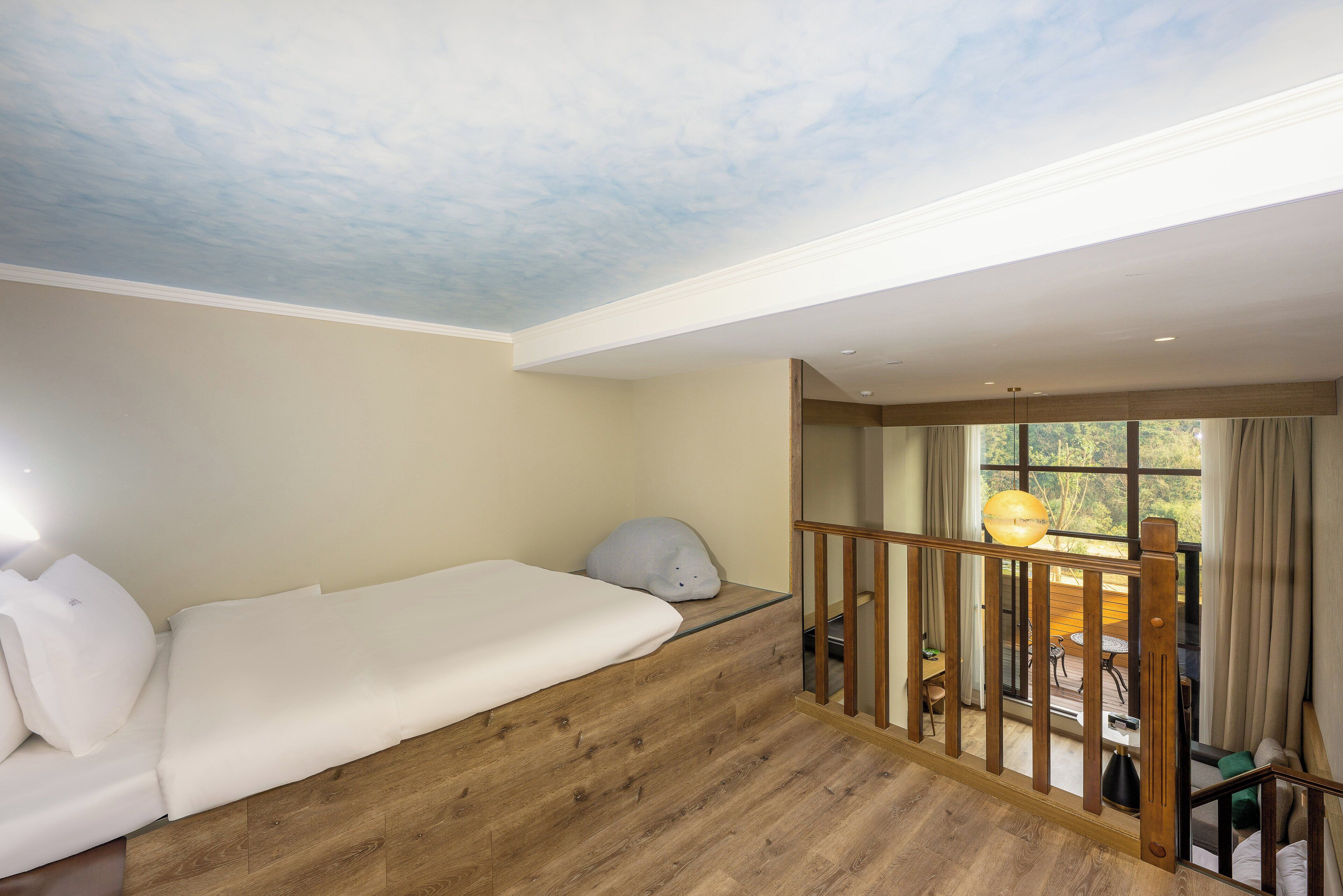 A room with a wooden floor, a white bed, a ceiling with a cloud design, and a view of an outdoor area with a table and chairs.