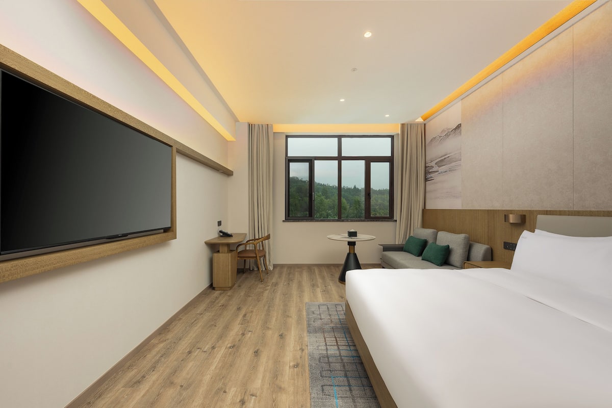 A hotel room with a large bed, a desk with a chair, a small table, and a large window with a view of greenery.