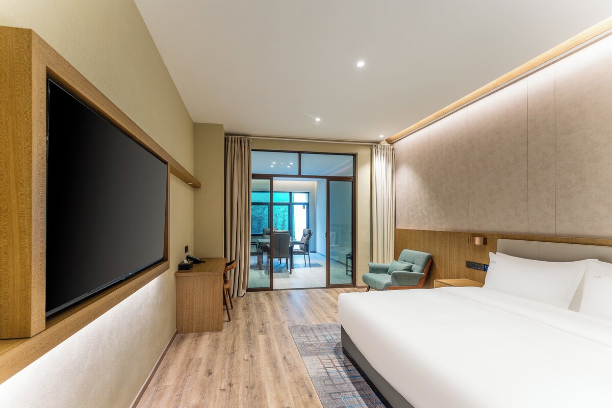 Una habitación de hotel moderna con una cama grande, un televisor de pantalla plana, un escritorio y vistas al exterior a través de una puerta corrediza de vidrio.