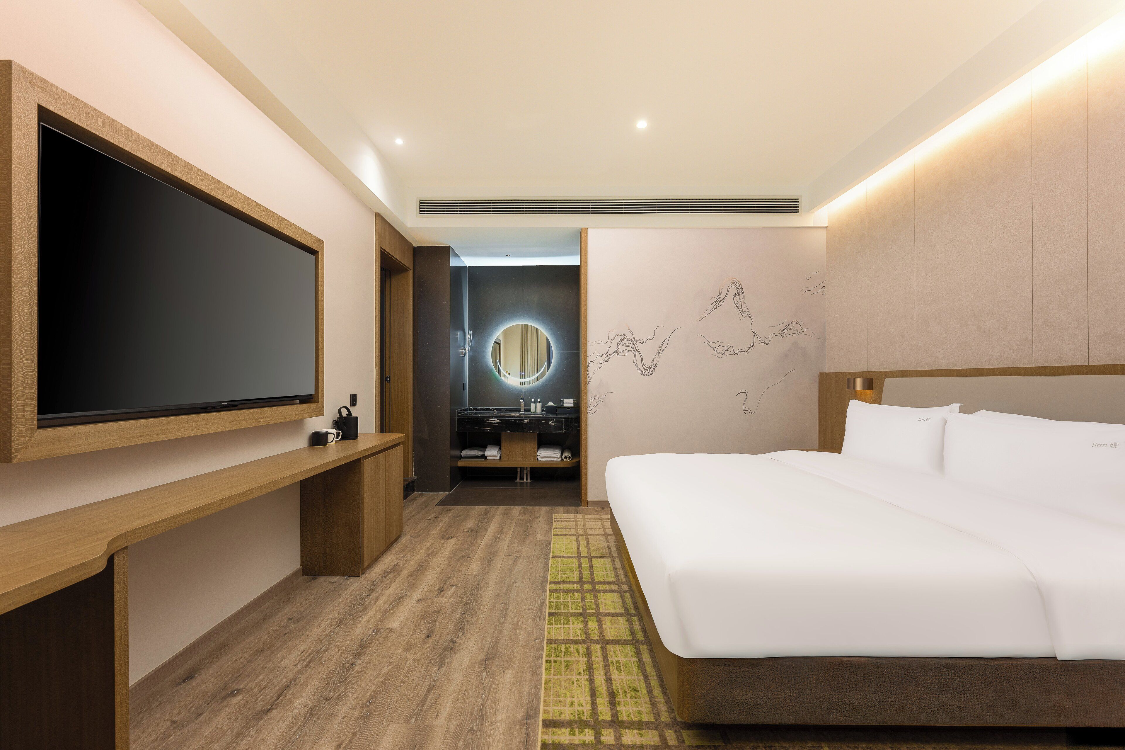 A modern hotel room with a large flat-screen TV, a wooden bench, a bed with white linens, a round mirror, and a wall with abstract art.