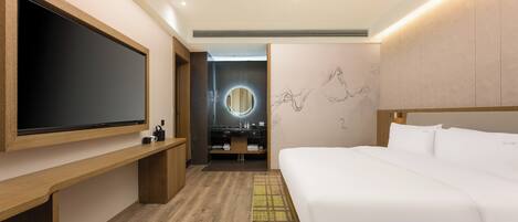 A modern hotel room with a large flat-screen TV, a wooden bench, a bed with white linens, a round mirror, and a wall with abstract art.