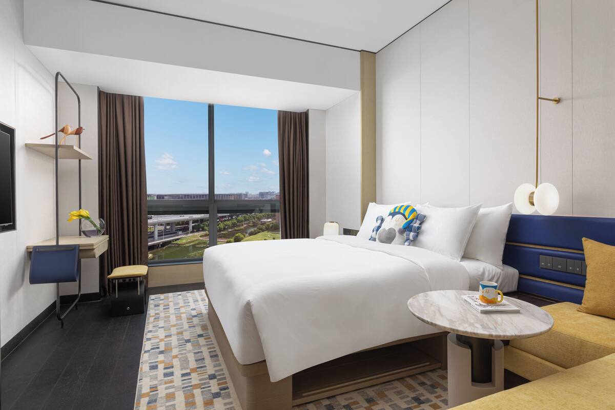 A modern hotel room with a large bed, a small desk, a chair, a round table, and a view of the city.