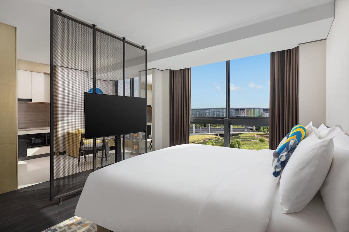 A hotel room with a large bed, a TV, a desk with a chair, and a view of a bridge and greenery.