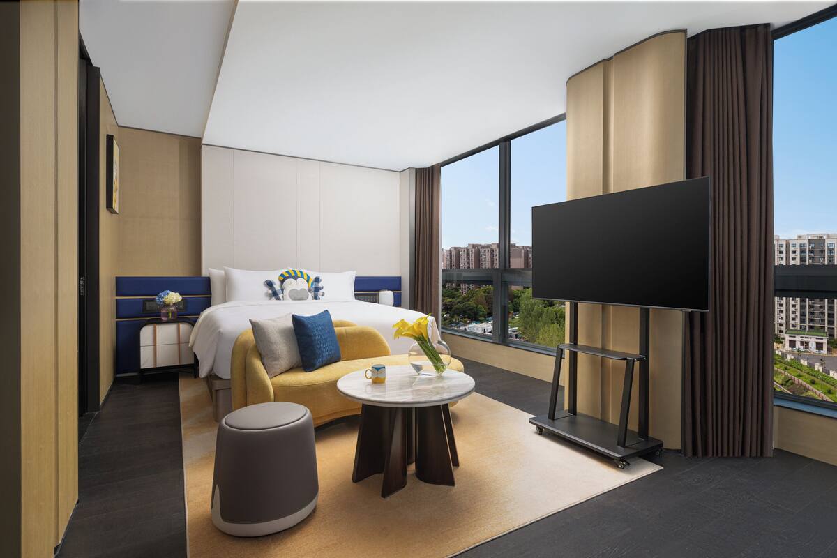 A modern hotel room with a bed, sofa, TV, and a view of the city.
