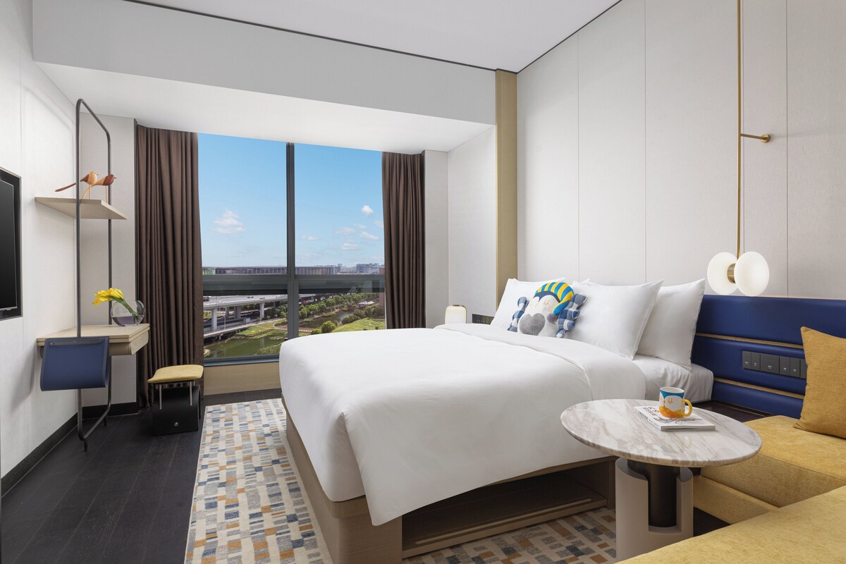 Habitación de hotel moderna con una cama grande, un escritorio pequeño, una silla, una mesa redonda y vistas a la ciudad.
