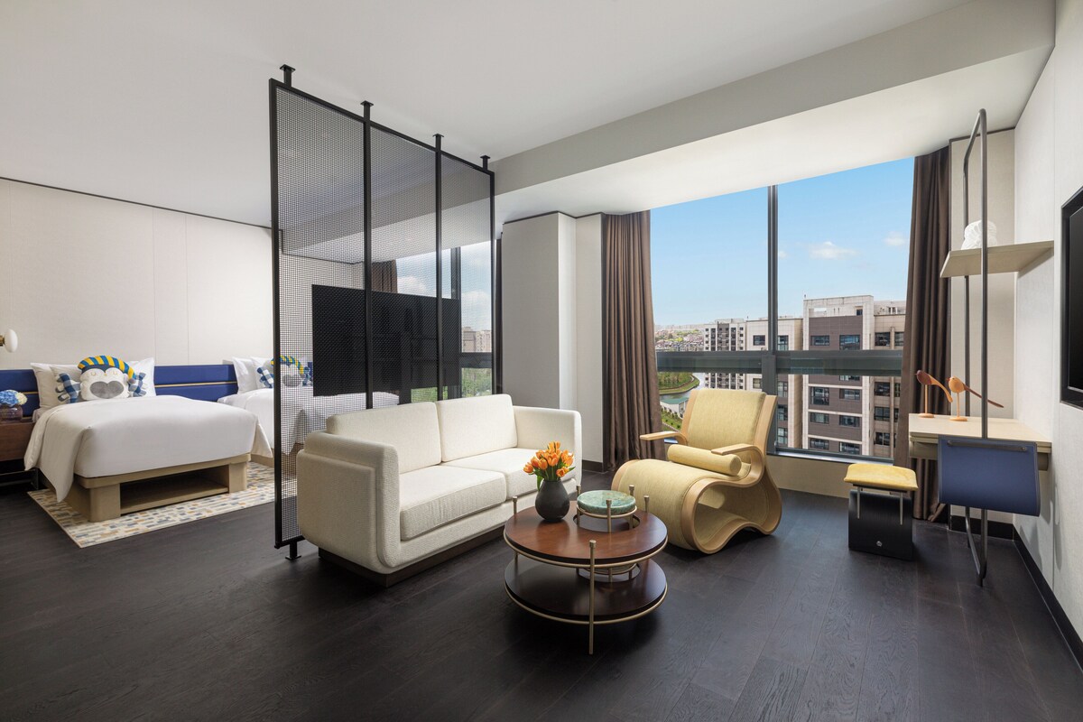 Una habitación de hotel moderna con cama, sofá, sillón, escritorio y un ventanal que ofrece vistas a la ciudad.