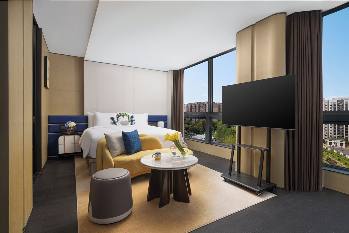 Habitación de hotel moderna con cama, sofá, televisor y vistas a la ciudad.