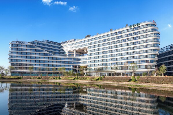 Un hôtel moderne à la façade incurvée, doté de nombreux balcons et dont l’image se reflète dans l’eau devant lui.