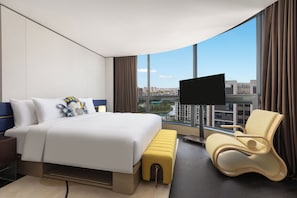 A modern hotel room with a large bed, a seating area, a television, and a view of the cityscape.