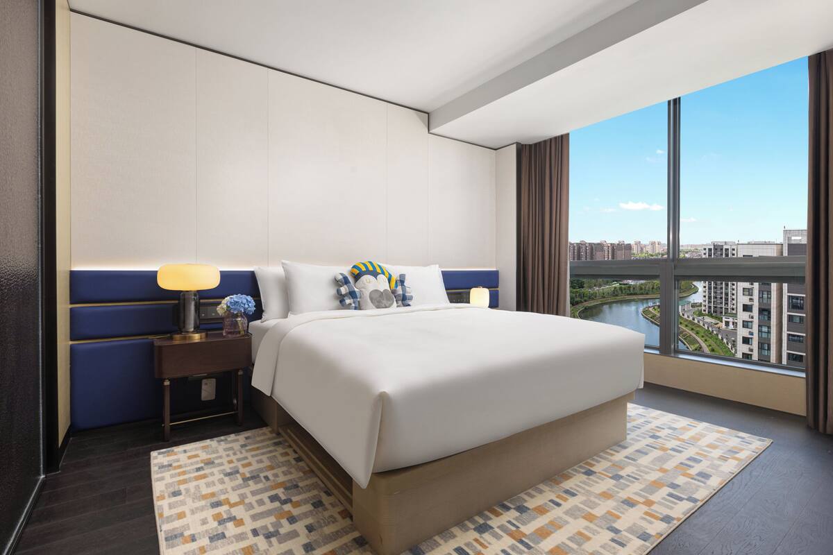 A modern hotel room with a large bed, a nightstand, a lamp, and a view of a cityscape through large windows.