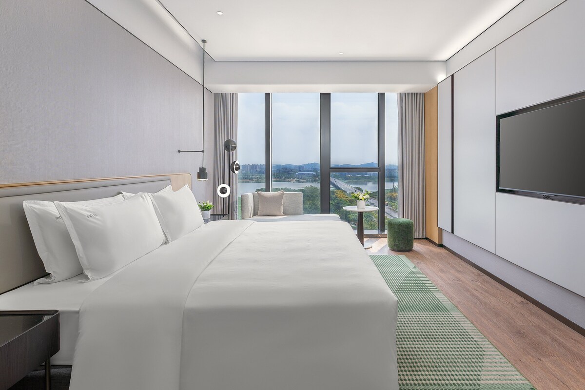 A modern hotel room with a large bed, a seating area, a TV, and a view of the city.
