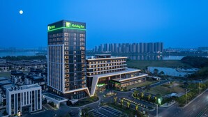 A modern hotel with a prominent 'Holiday Inn' sign, multiple floors, and a view of a cityscape and water body in the background. - Holiday Inn Wuhan Houguan Lake by IHG (Wuhan)