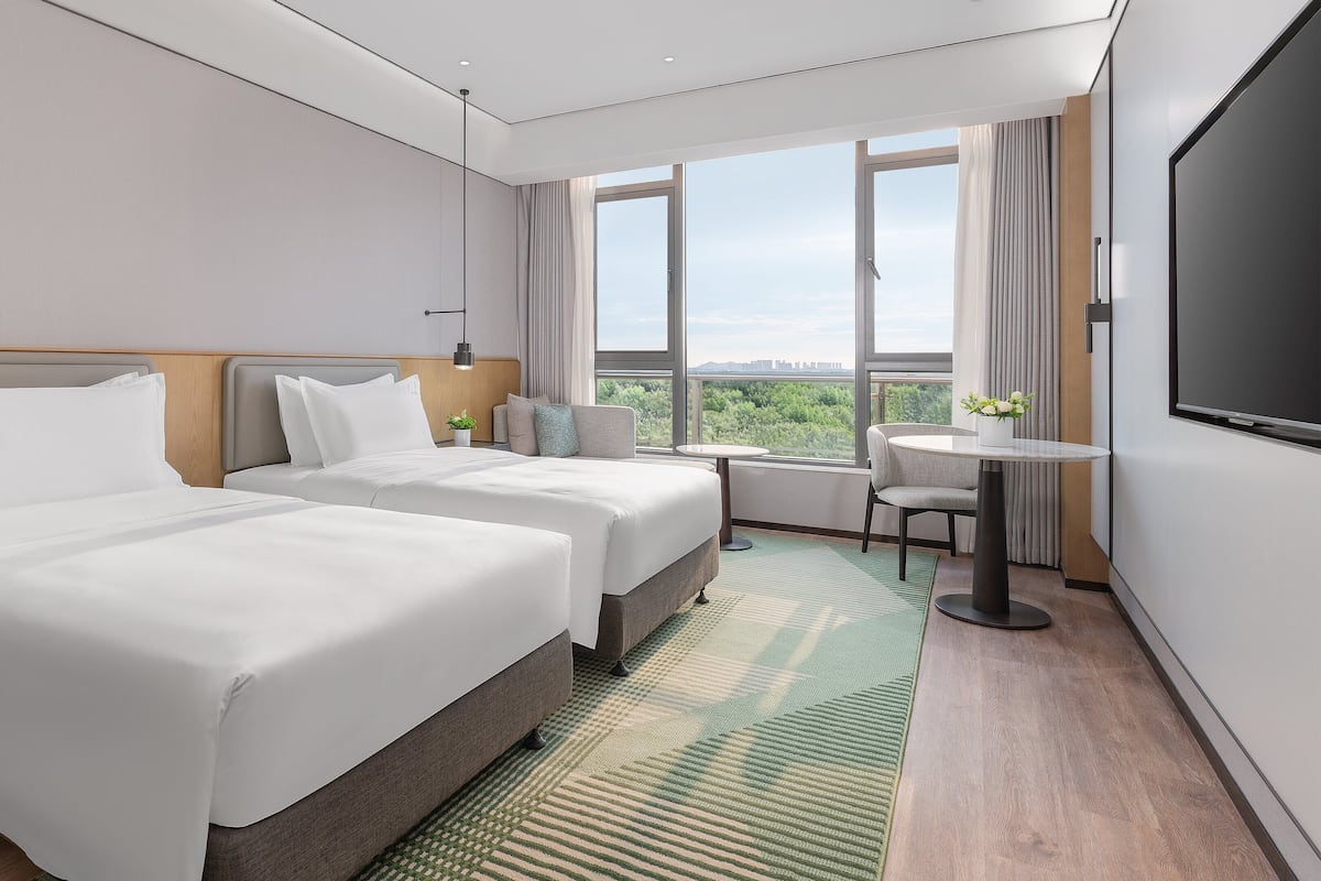 A hotel room with two beds, a TV, a small table, and a view of the city.