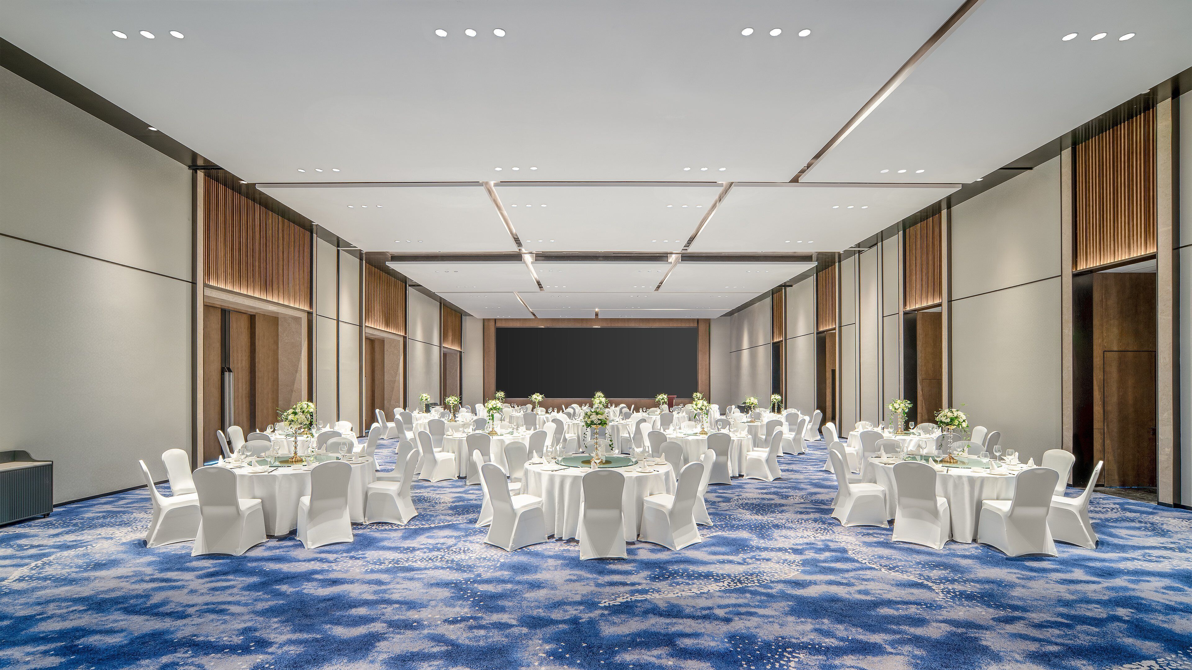 Un amplio salón de conferencias bien iluminado, con filas de mesas y sillas blancas, una alfombra azul y una gran pantalla al frente.