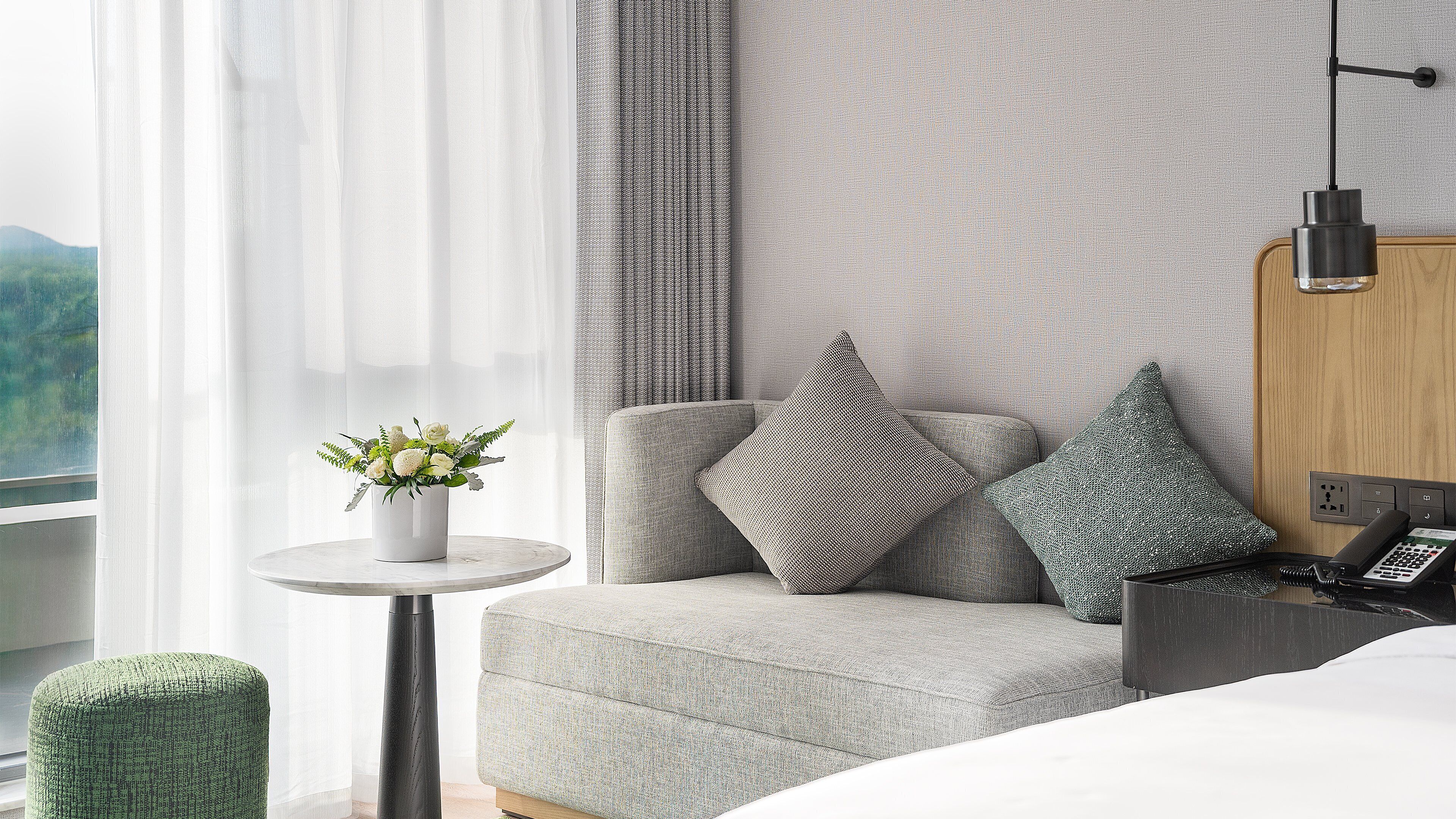 A modern hotel room with a grey sofa, two patterned pillows, a small white table with a plant, and a bedside table with a phone and remote control.