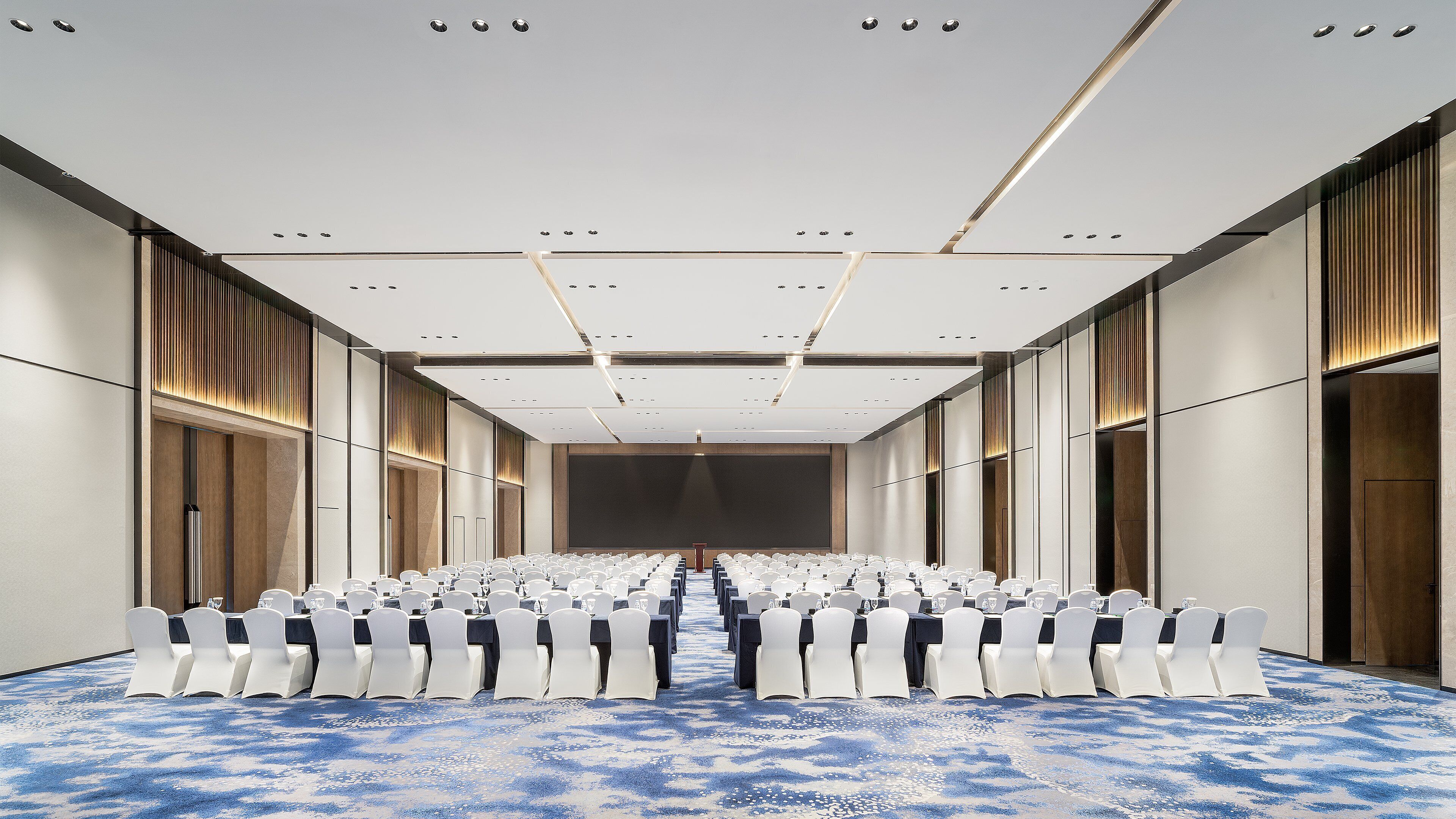 A conference room with rows of white chairs, a large screen at the front, and a carpeted floor.