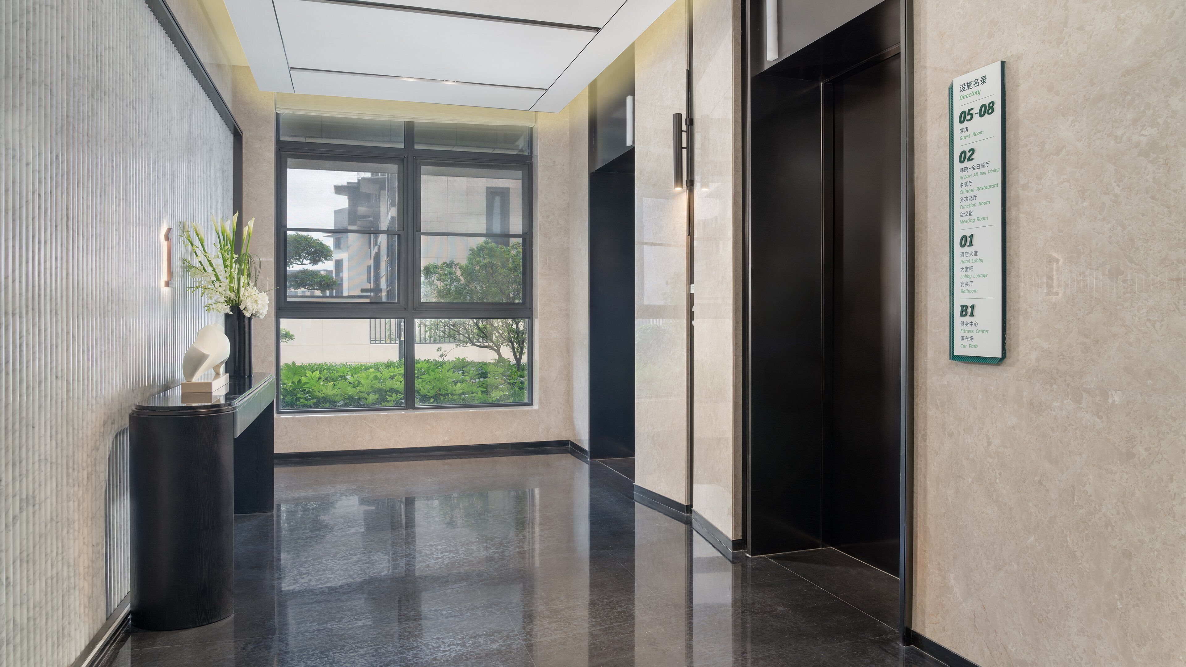 Hall d'hotel moderna con un ascensore in vetro, una segnaletica direzionale e vista sull'esterno.