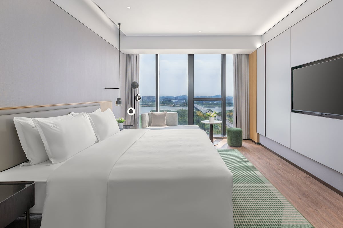 A modern hotel room with a large bed, a seating area, a TV, and a view of the city.