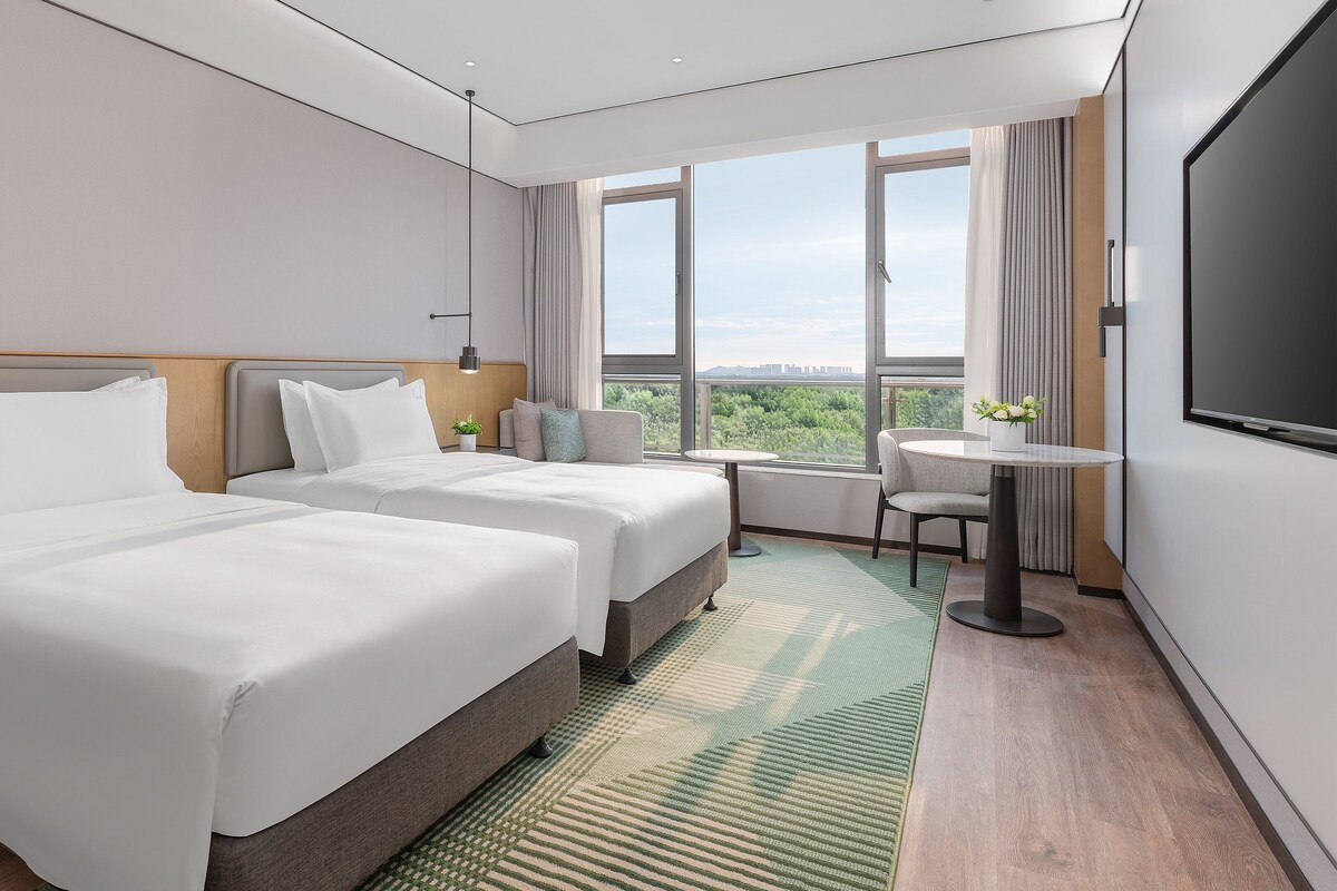 A hotel room with two beds, a TV, a small table, and a view of the city.