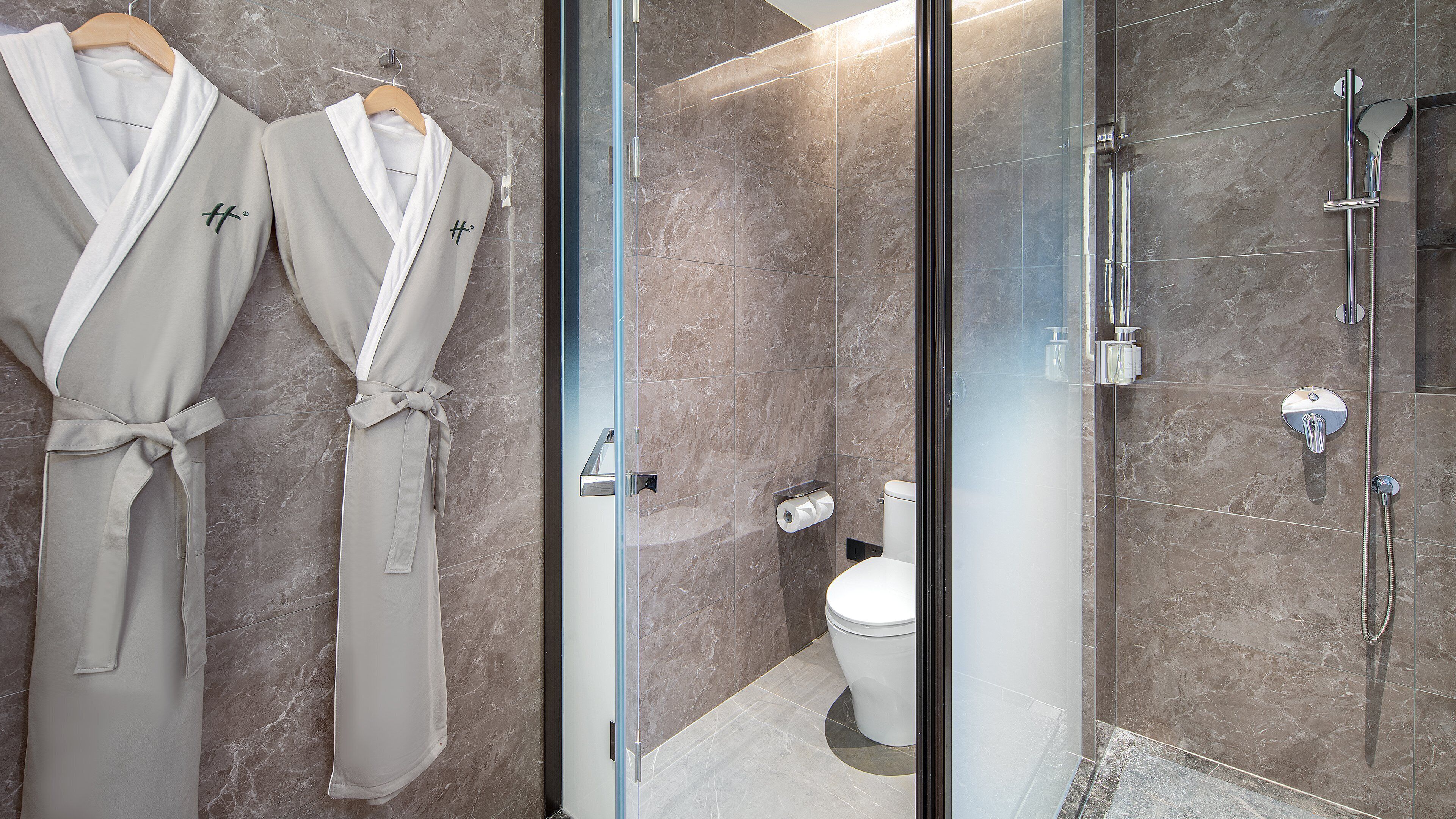 A bathroom with a glass shower enclosure, a toilet, and two robes hanging on a rack.