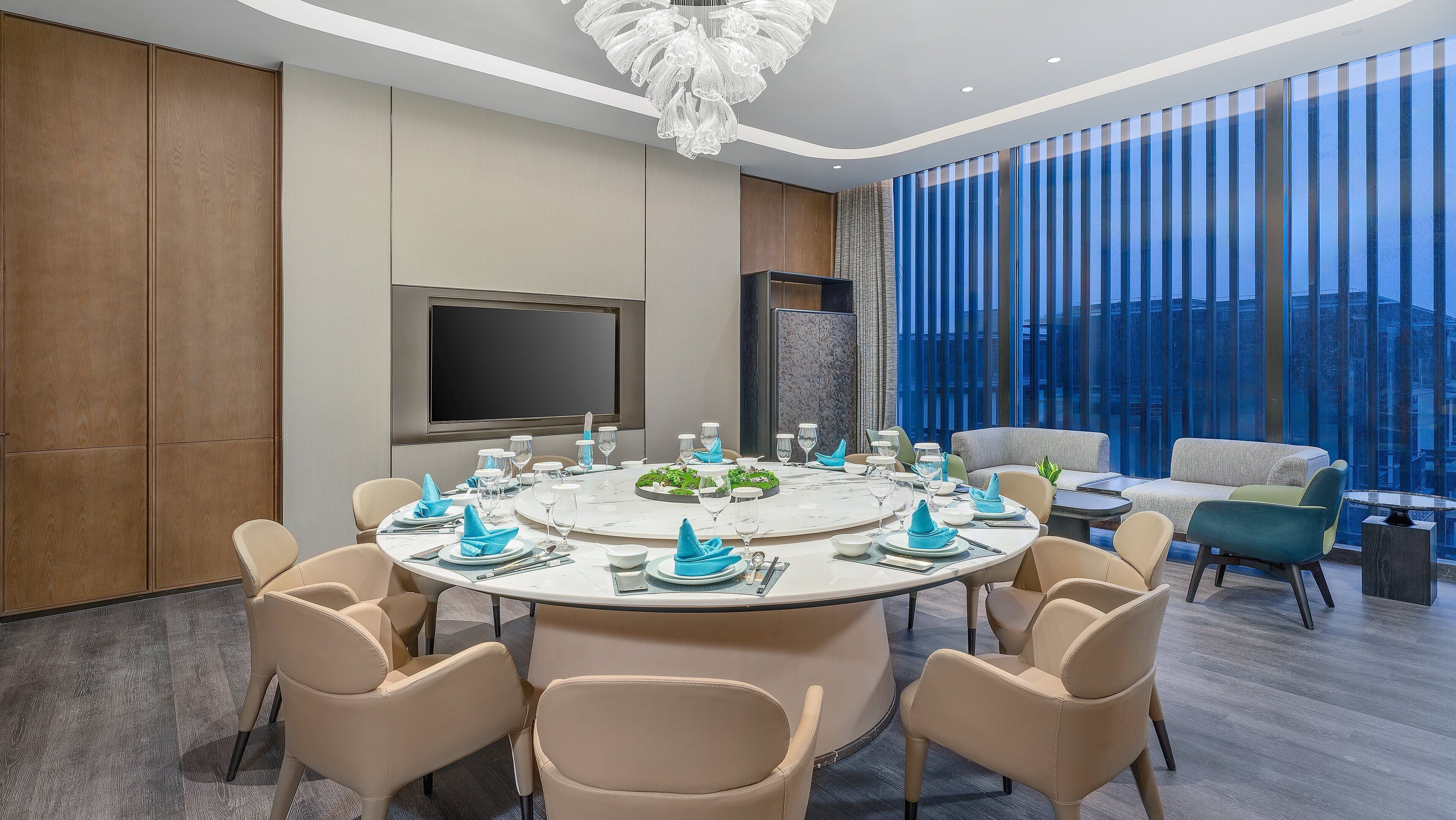 Un comedor moderno con una mesa redonda dispuesta para seis personas, rodeada de sillas color beige. Un televisor de pantalla plana grande está montado en la pared, y la habitación tiene un ventanal con cortinas azules.
