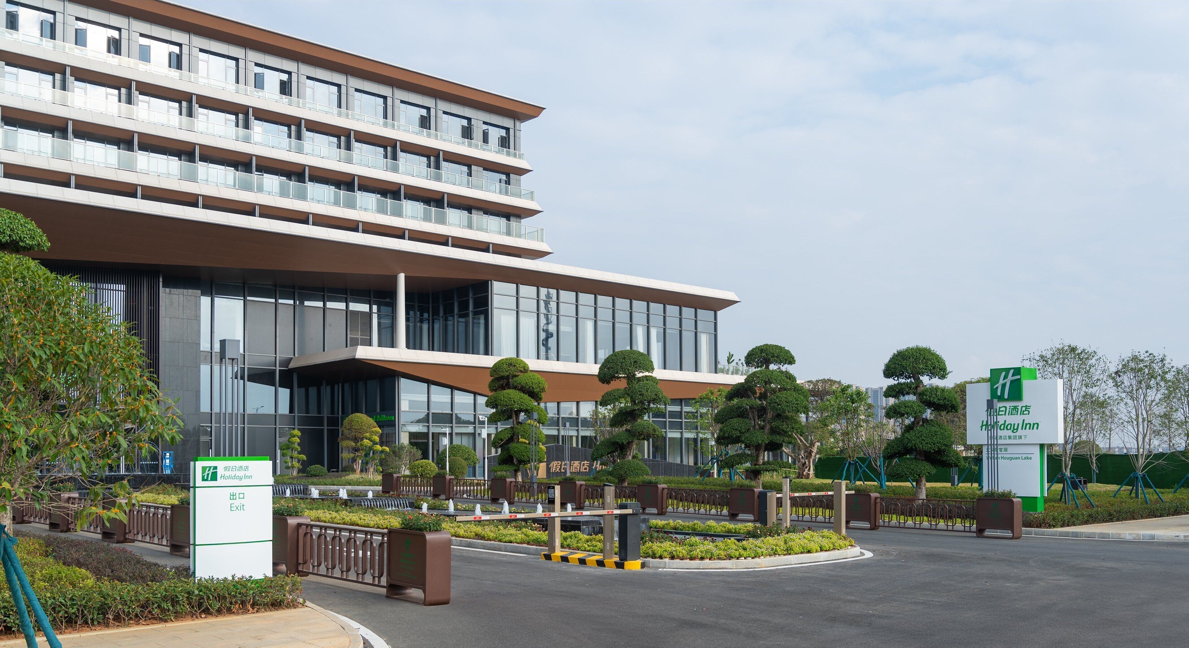 Un hôtel moderne, avec plusieurs étages, de grandes fenêtres et un jardin bien entretenu, agrémenté d’arbres et d’arbustes aux formes soignées.
