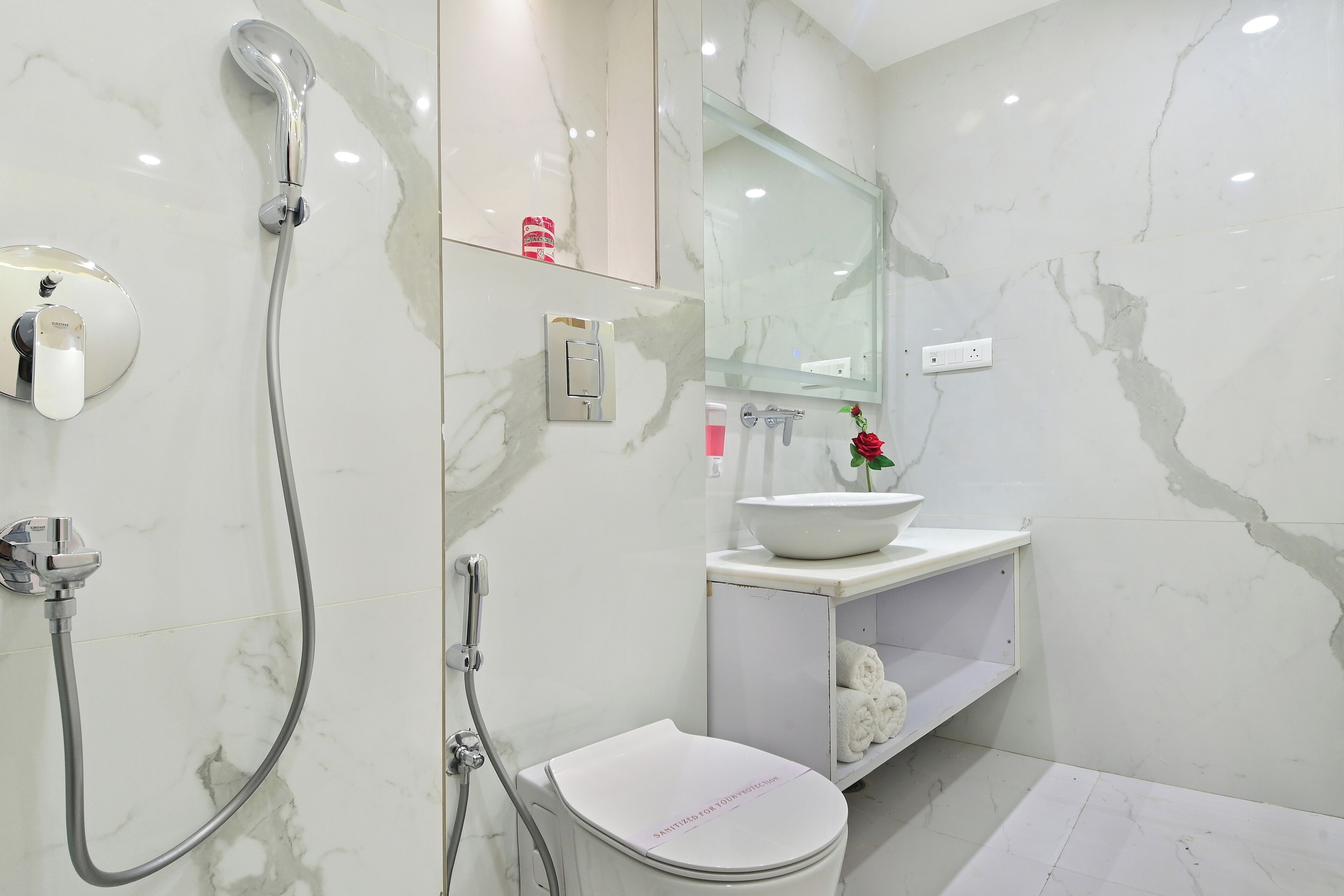 Une salle de bain moderne avec un sol en marbre blanc, des toilettes blanches, un lavabo agrémenté d’un vase de fleurs et une douche équipée d’un pommeau de douche à main.
