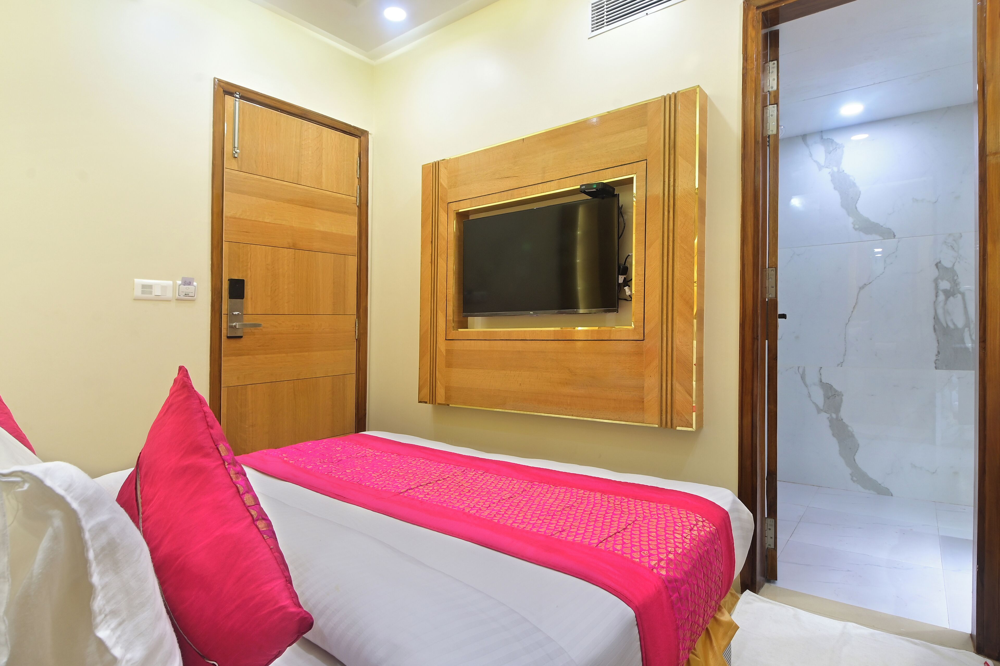 A hotel room with a bed, a wooden door, a television mounted on the wall, and a bathroom with a glass door.