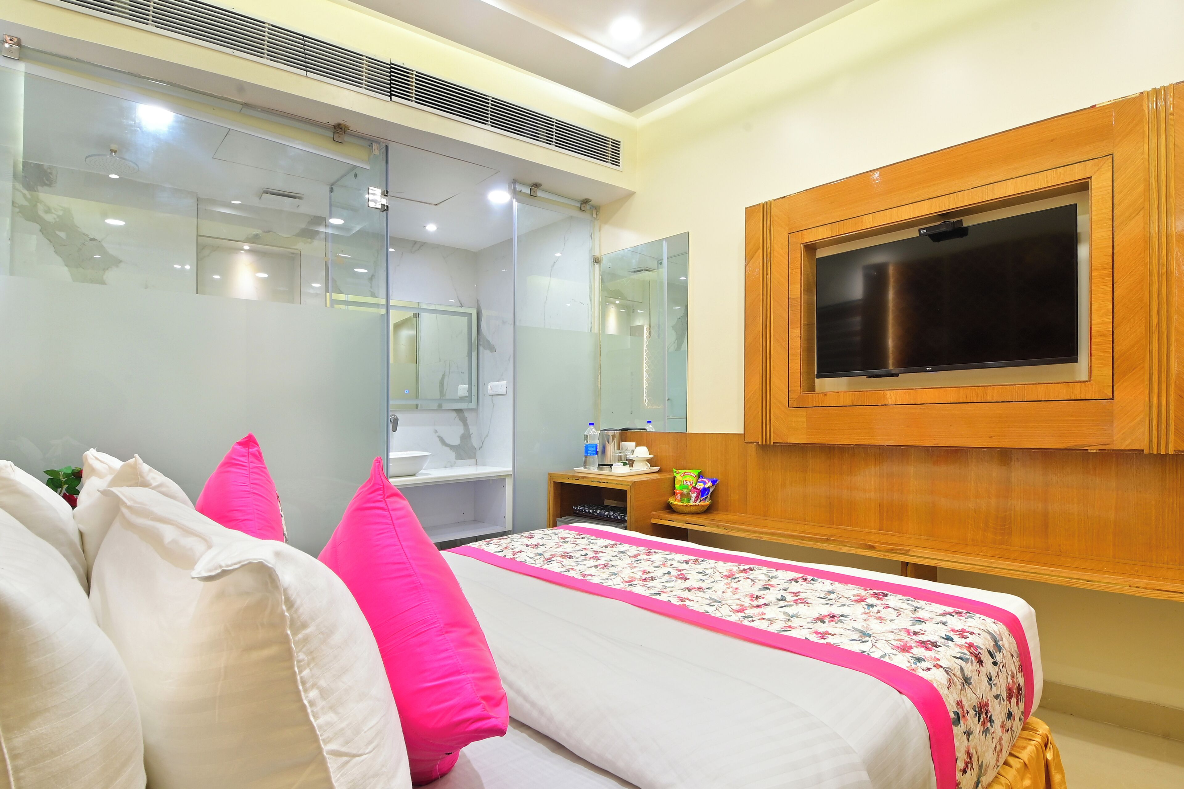 Habitación de hotel con cama, televisor montado en la pared, espejo y baño visible a través de un mamparo de vidrio.