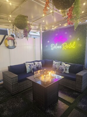 A cozy outdoor seating area with a neon sign, a fire pit, and decorative plants.