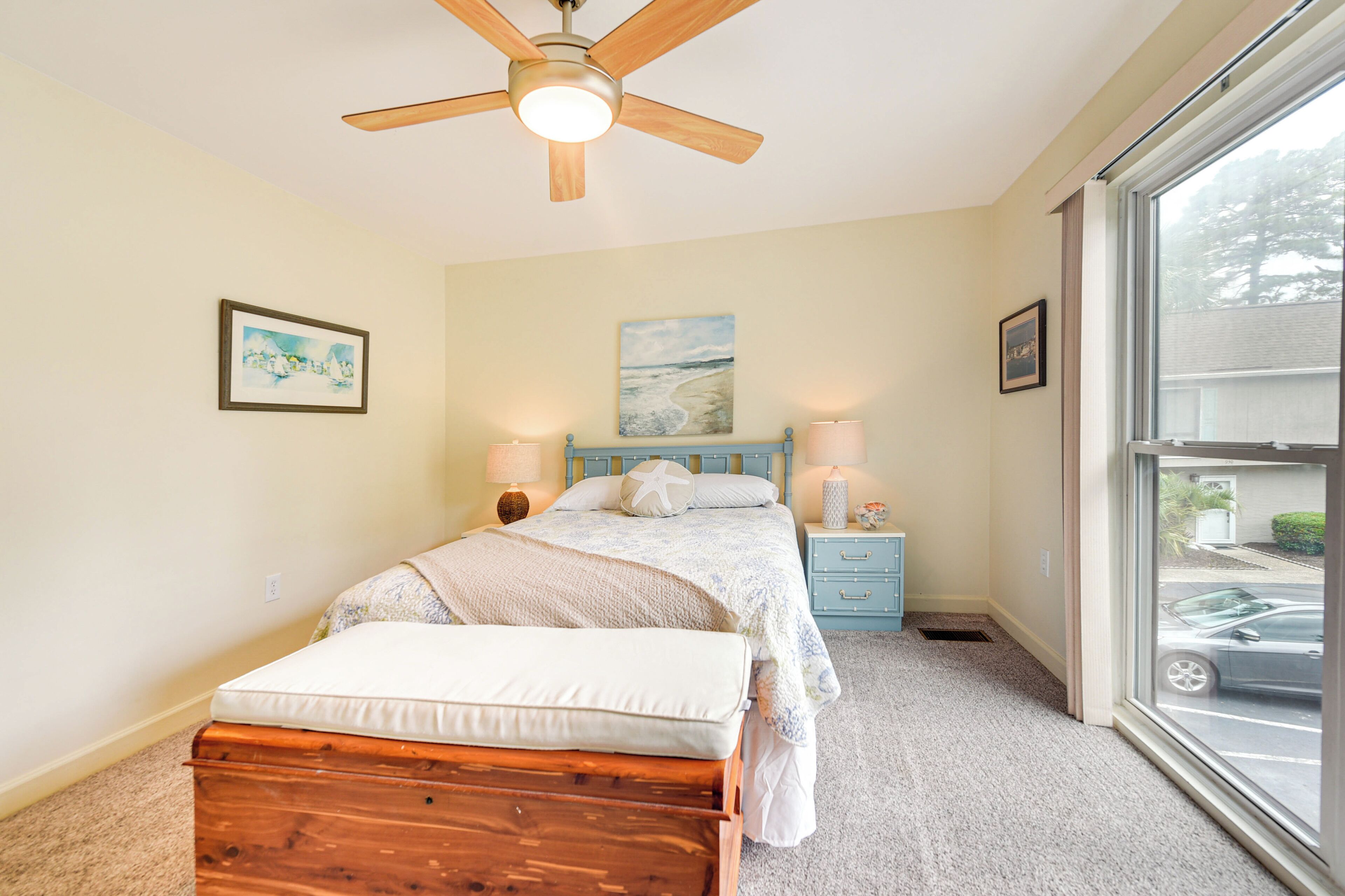 A bedroom with a bed, a nightstand, a ceiling fan, and a window with a view of a parking lot.