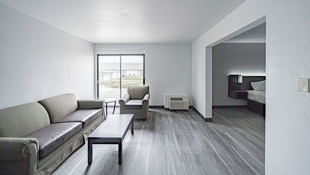 A modern hotel room with a grey sofa, a small table, and a chair. There is a bed with a headboard and a bedside table. The room has a large window and an air conditioning unit. - Studio 6 Pocatello, Id (Pocatello)