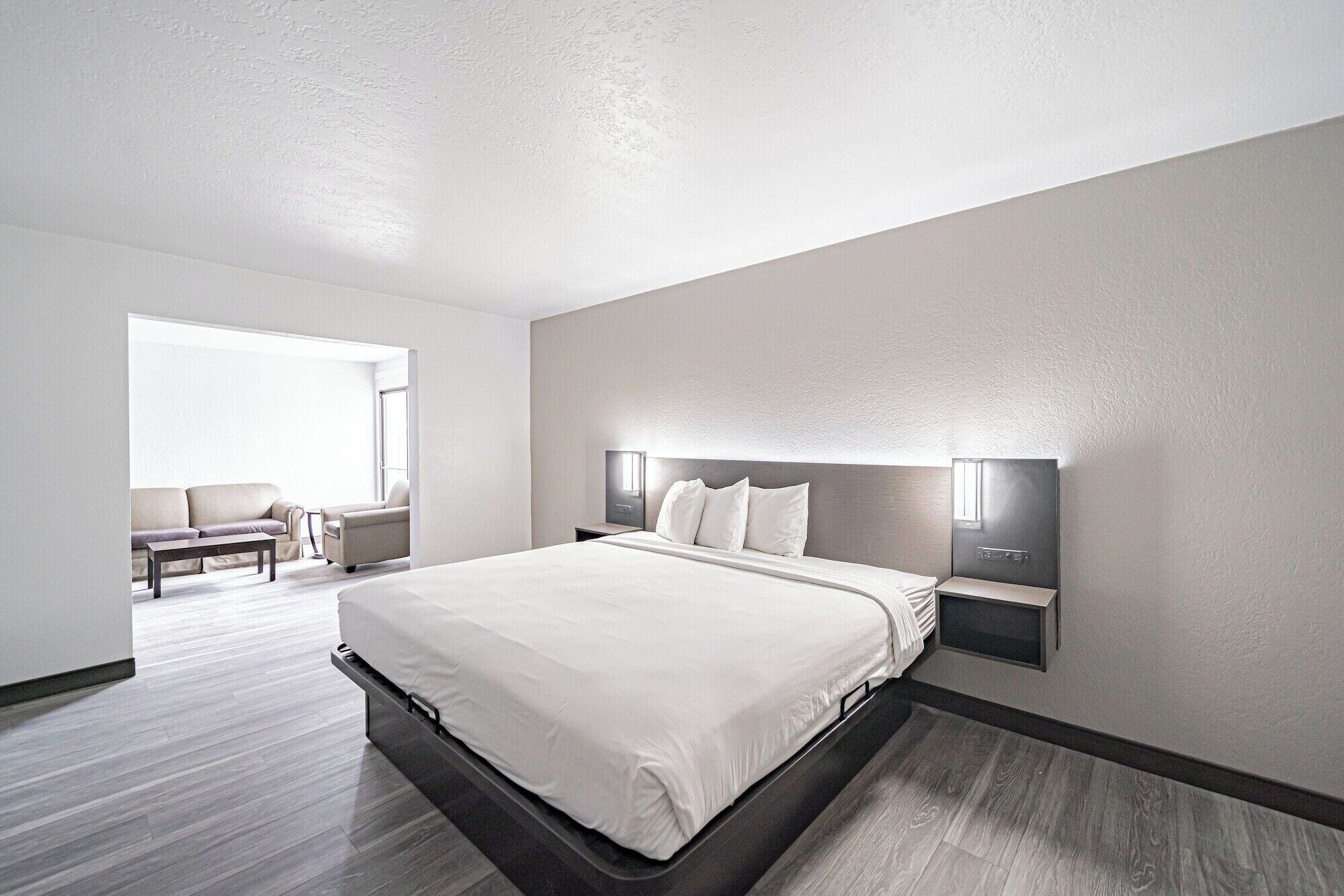 A modern hotel room with a large bed, a seating area with a sofa and armchairs, and a large window providing natural light.