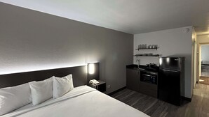 A hotel room with a large bed, a bedside table with a phone, a small kitchenette with a stove and refrigerator, and a wall-mounted shelf with cups. - Studio 6 Pocatello, Id (Pocatello)