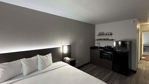 A hotel room with a large bed, a bedside table with a phone, a small kitchenette with a stove and refrigerator, and a wall-mounted shelf with cups. - Studio 6 Pocatello, Id (Pocatello)