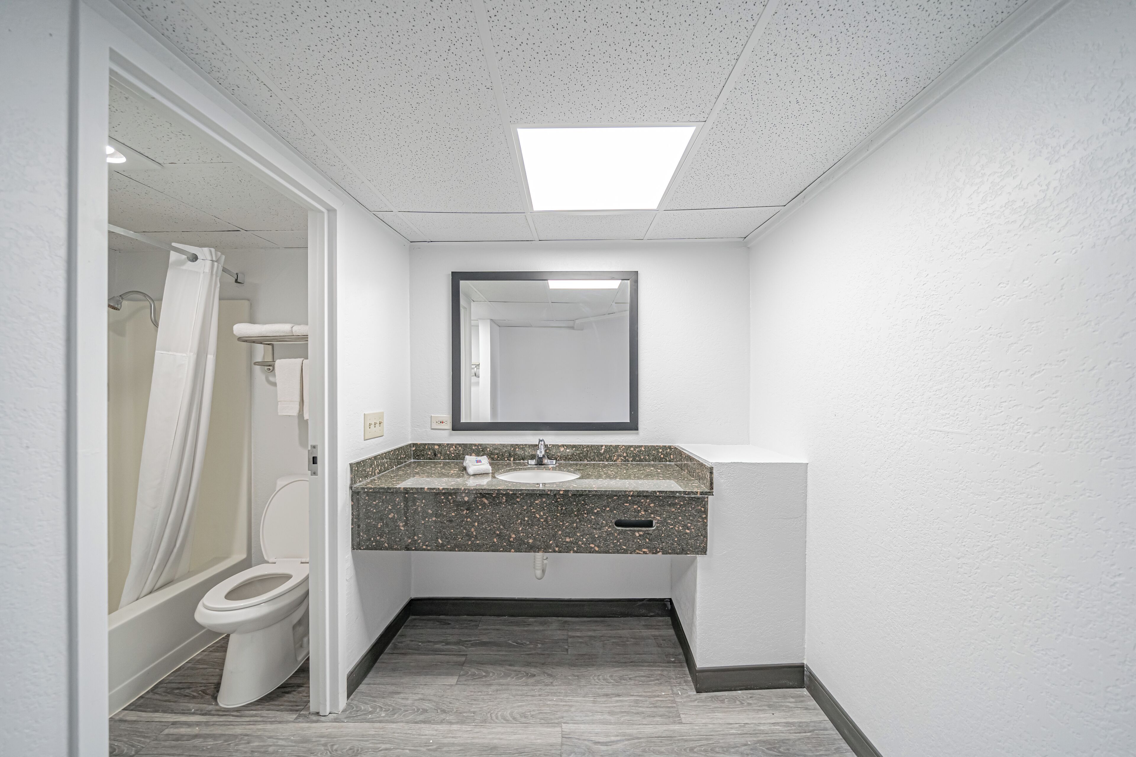 A bathroom with a white toilet, a shower with a white curtain, a granite countertop sink, and a mirror above it.