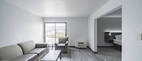 A modern, minimalist hotel room with a grey sofa, a small table, and a chair. There is a large window providing a view of the outside.