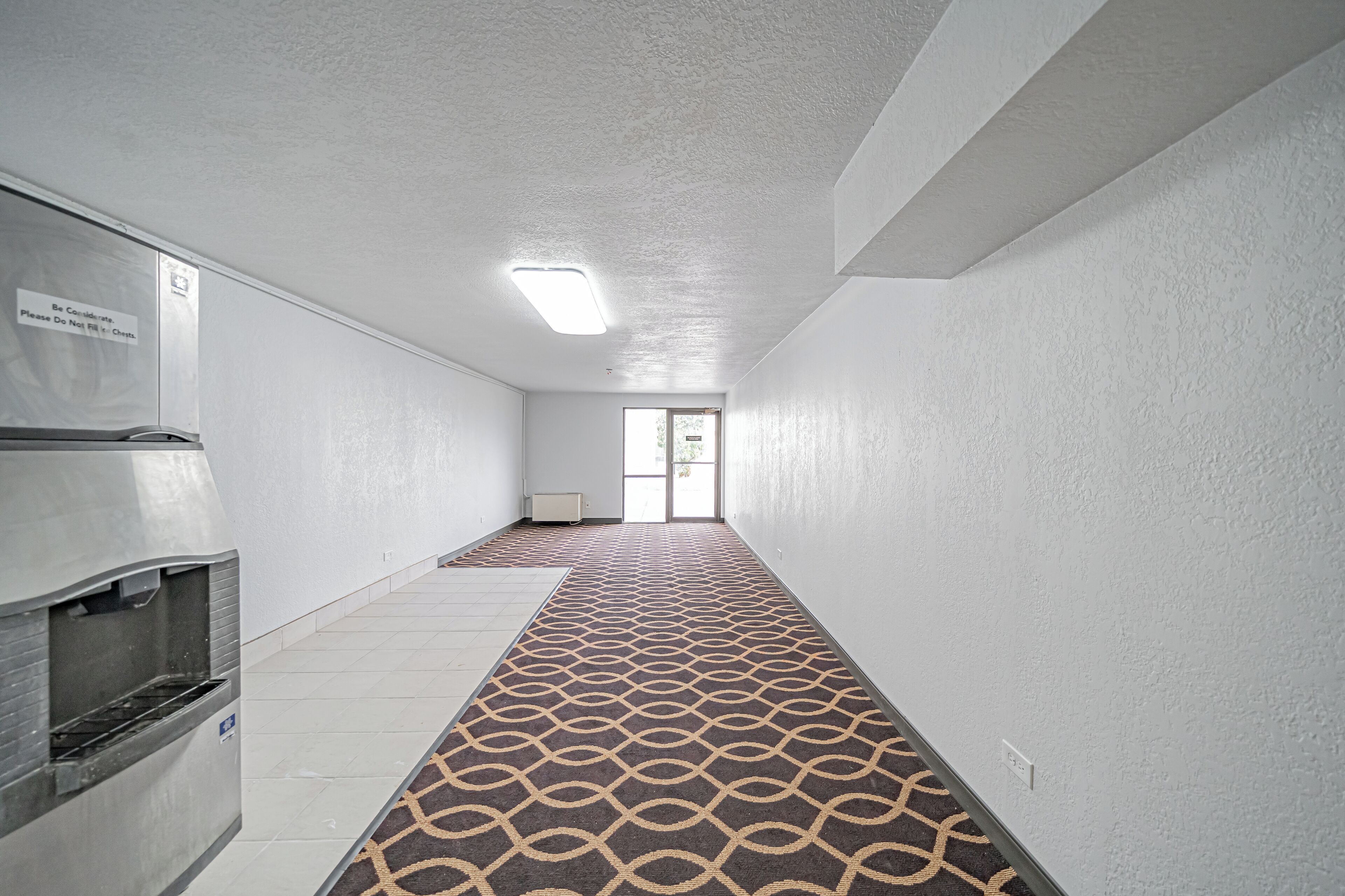 A hallway with a patterned carpet, a refrigerator, and a door leading to a bright room.